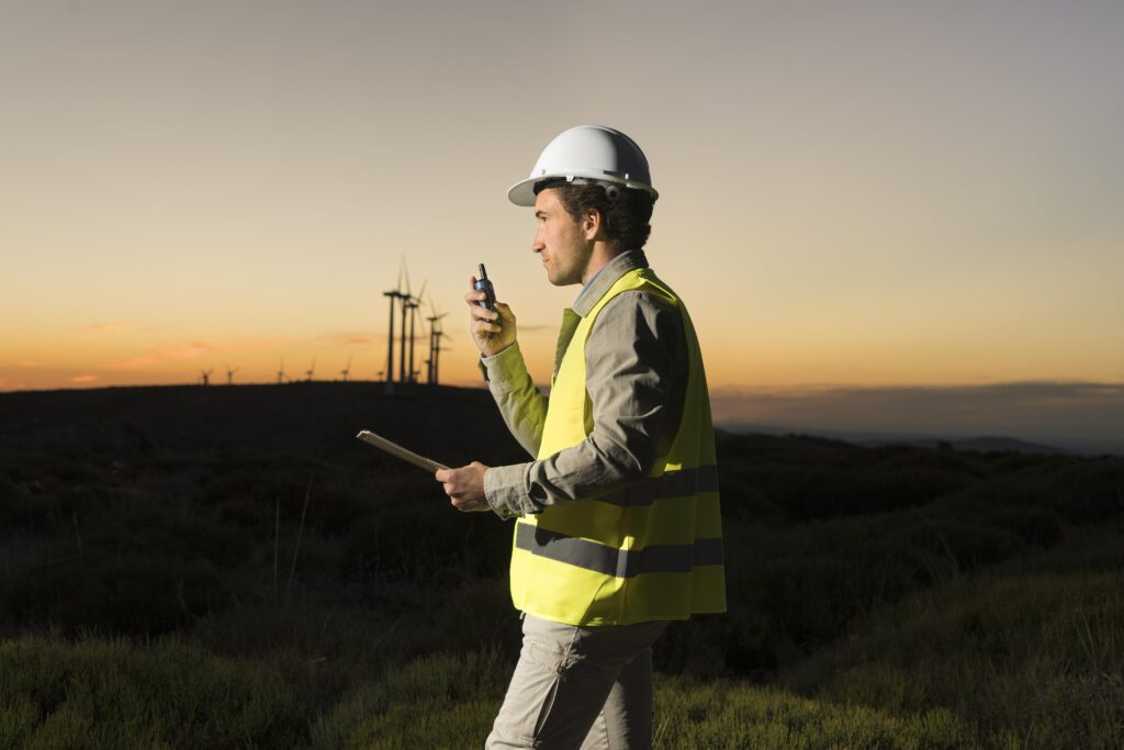Image of a renewable energy technician in a hard hat and safety vest, holding a radio and clipboard, standing near a field of wind turbines at sunset